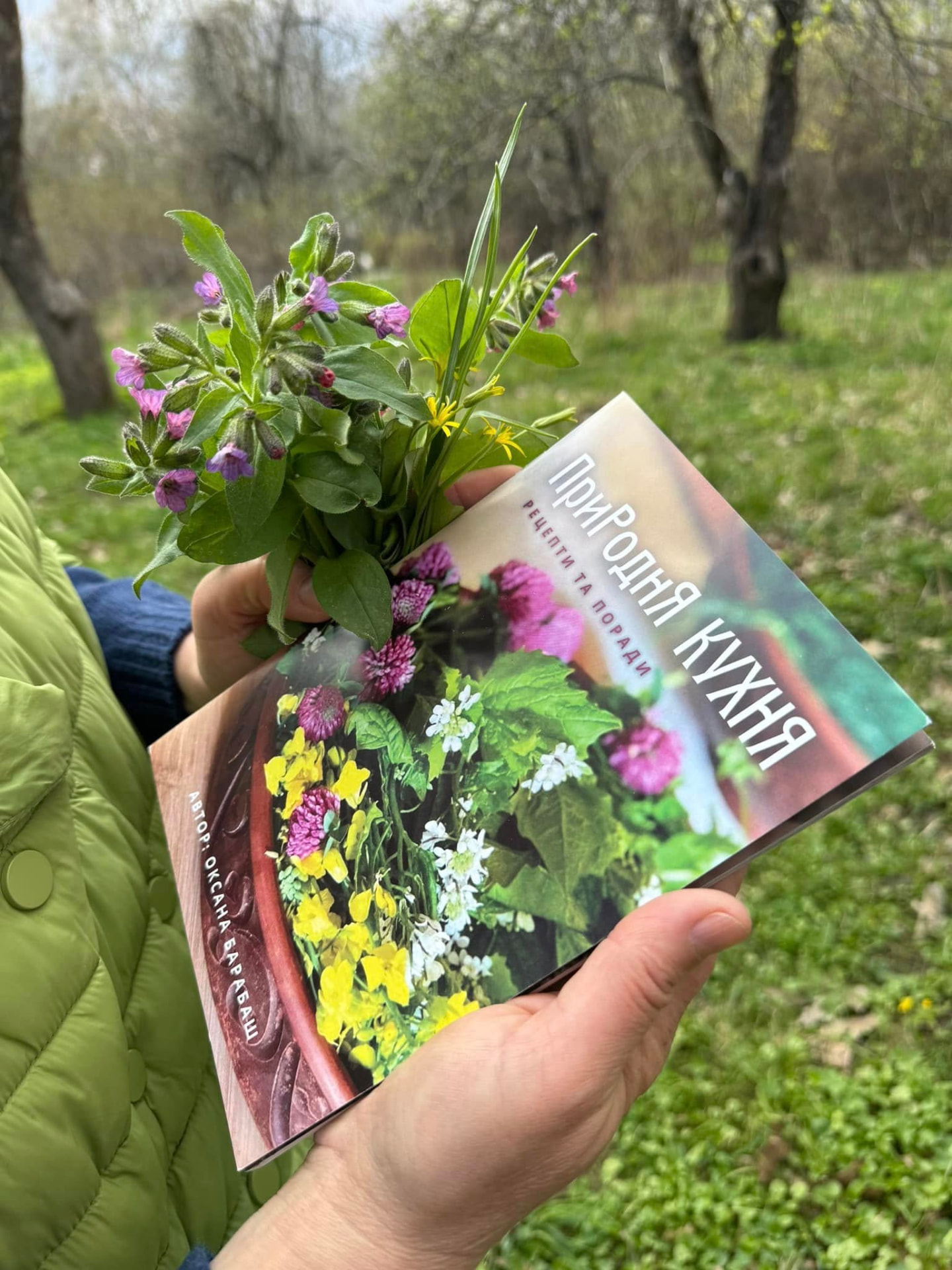 Книга «ПриРоднЯ КУХНЯ. Рецепти та поради» Оксани Барабаш у руках з букетом весняних дикоросів